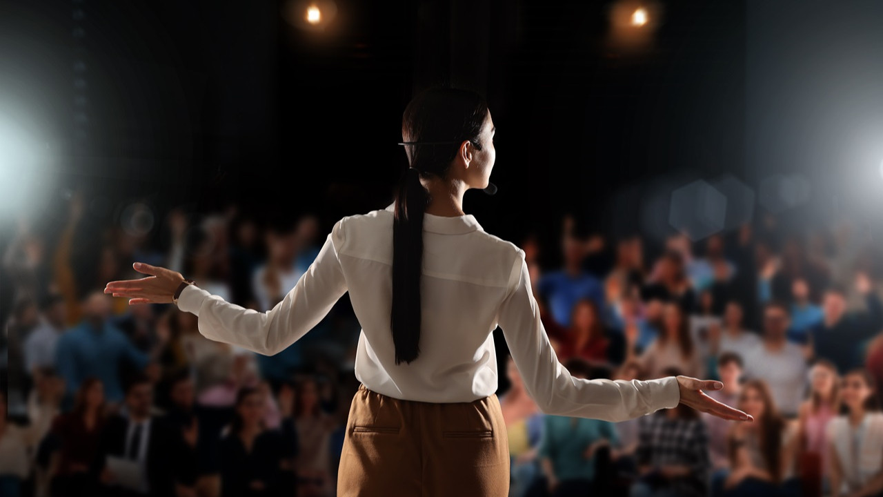 Səhnədə auditoriyaya nitq söyləyən qadın natiq. Ağ paltar geyinmiş, uzun saçlı qadın əllərini hərəkət etdirərək insanlara xitab edir.